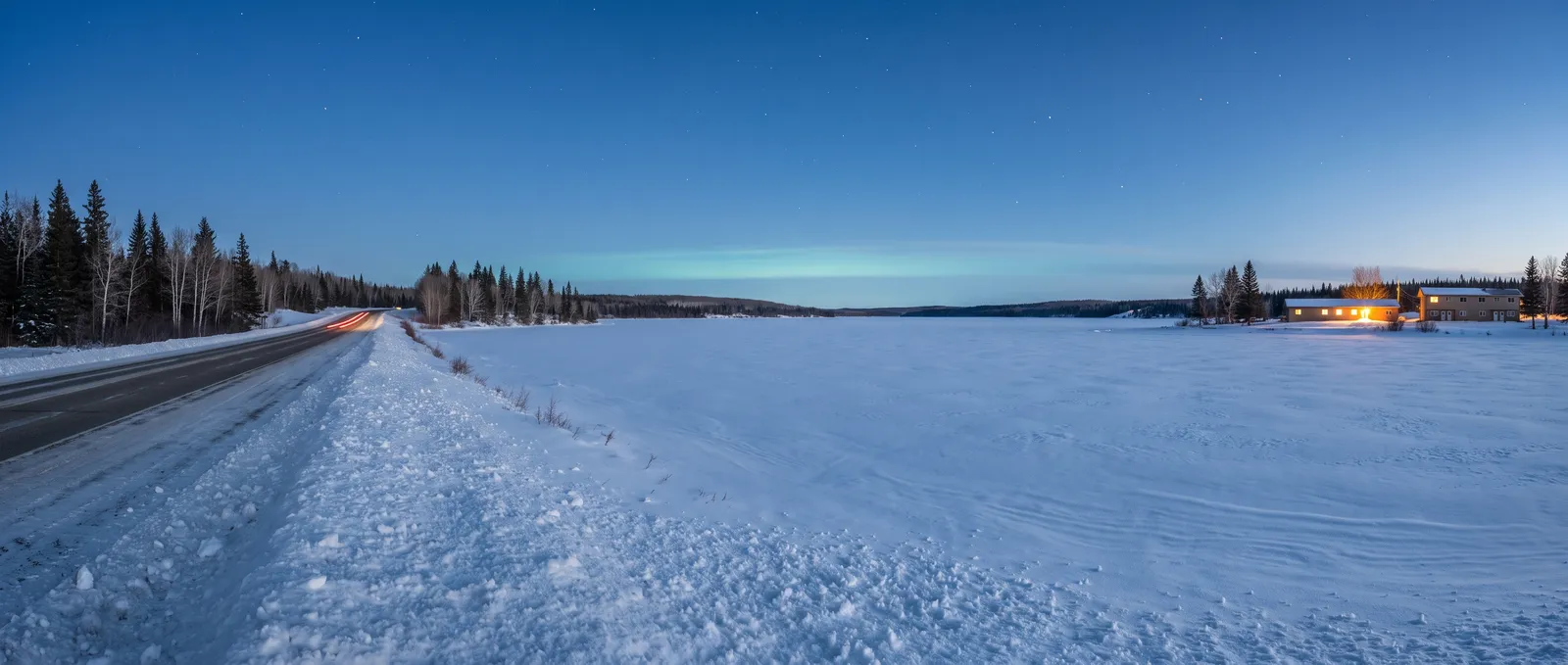 Generated scene: a snow-covered frozen lake at blue hour, a two-lane highway curving along the shore between snowbanks, warm lit windows of low buildings far across the snow and the first stars.
