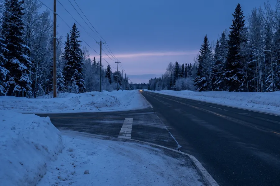 Generated scene: a two-lane highway through snowy spruce and aspen forest at dusk, power poles along the road and one pair of headlights far away.
